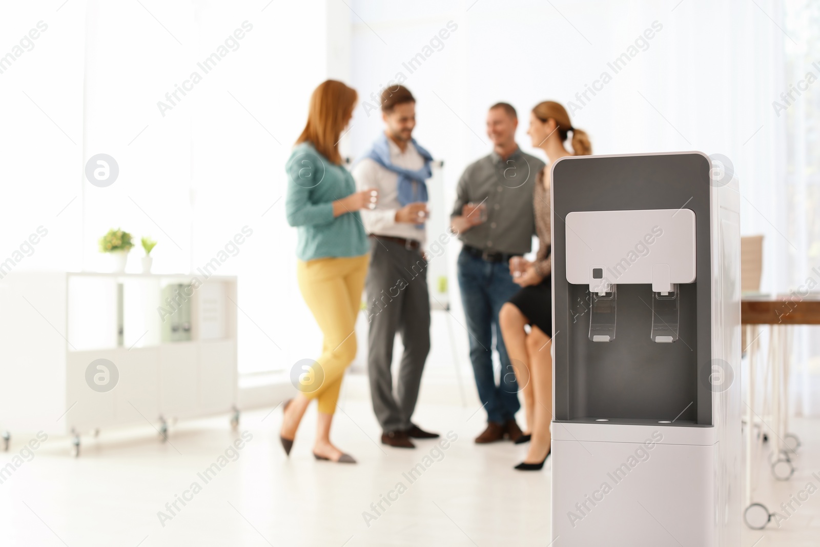 Photo of Modern water cooler and blurred office employees on background. Space for text