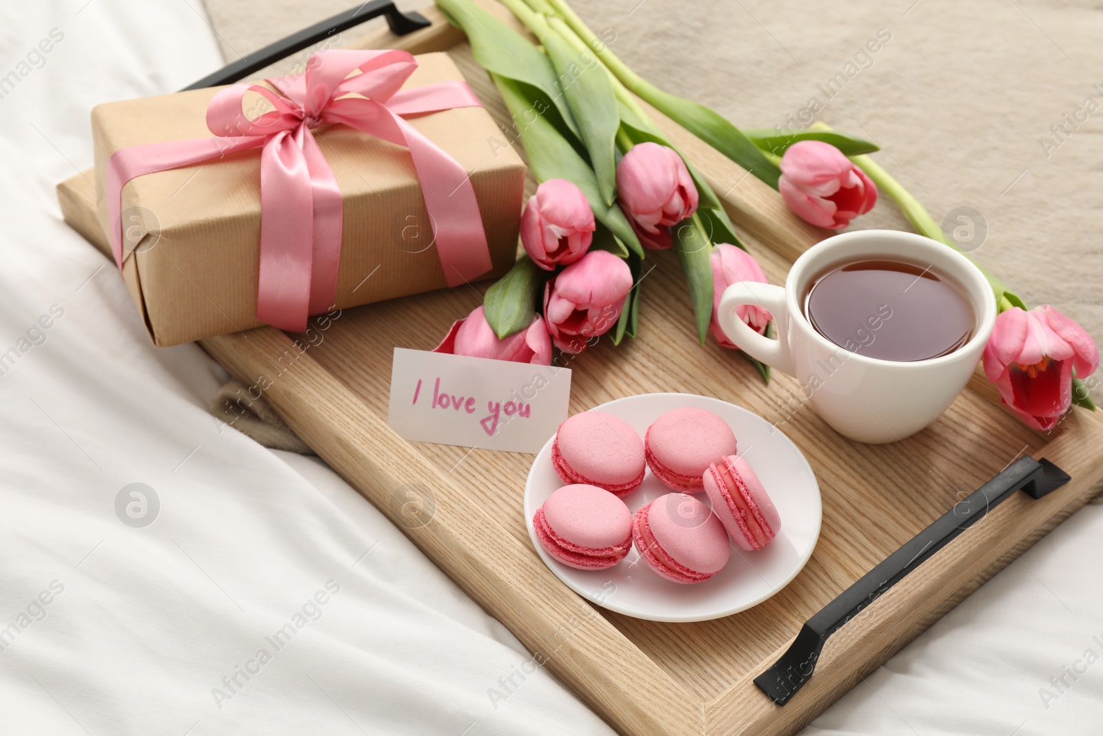 Photo of Tasty breakfast served in bed. Delicious macarons, tea, flowers, gift box and card with phrase I Love You on tray