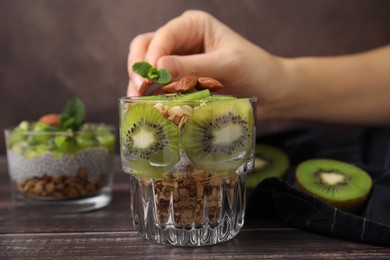 Woman putting mint on delicious dessert with kiwi, muesli and almonds at wooden table, closeup