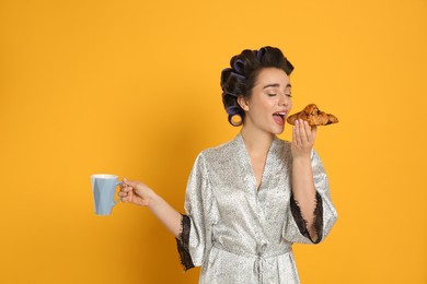 Photo of Young woman in silk bathrobe with hair curlers having breakfast on orange background