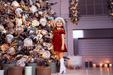 Photo of Cute little child wearing Santa hat near Christmas tree at home
