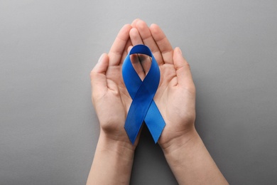 Woman holding blue awareness ribbon on grey background, top view