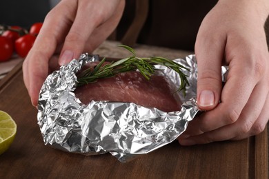 Woman wrapping meat with rosemary in aluminum foil at wooden table, closeup