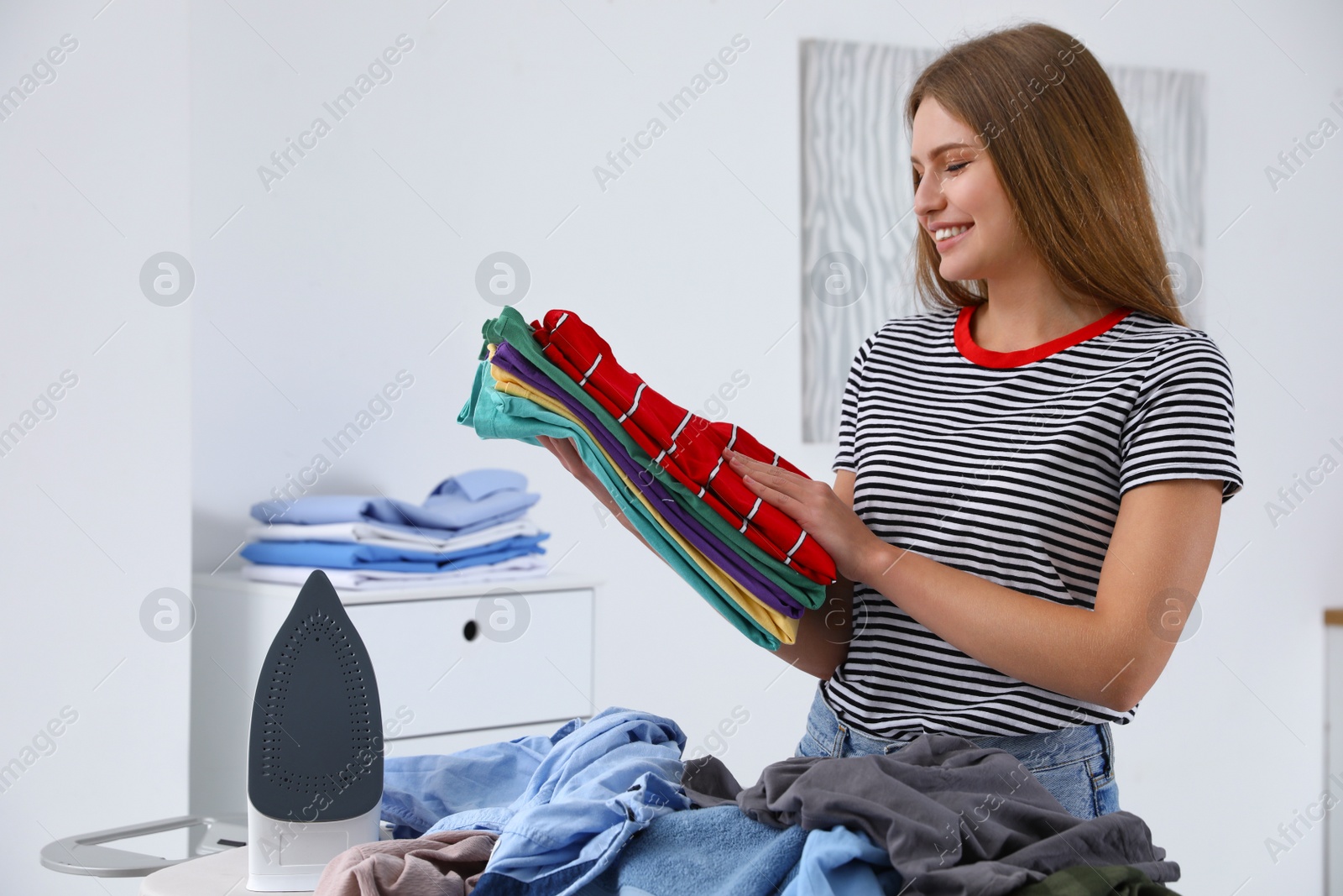 Photo of Young woman with folded clothes near ironing board at home