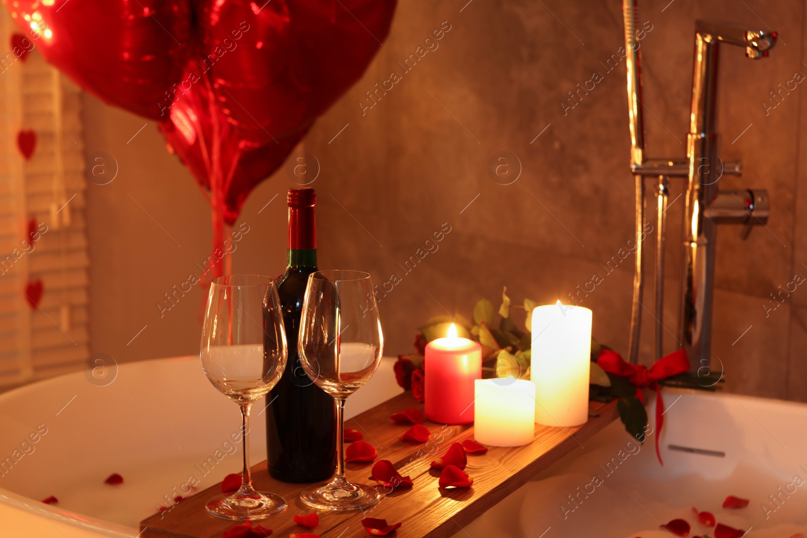 Photo of Wooden tray with wine, burning candles and rose petals on tub in bathroom. Valentine's day celebration