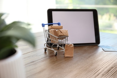 Photo of Internet shopping. Small cart with boxes and bag near modern tablet on wooden table indoors