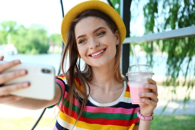 Photo of Happy young woman with drink taking selfie in park