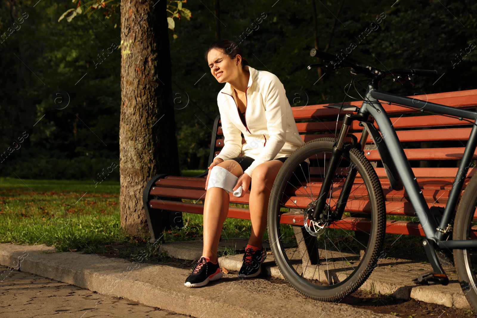 Photo of Young woman applying bandage onto her knee on wooden bench outdoors