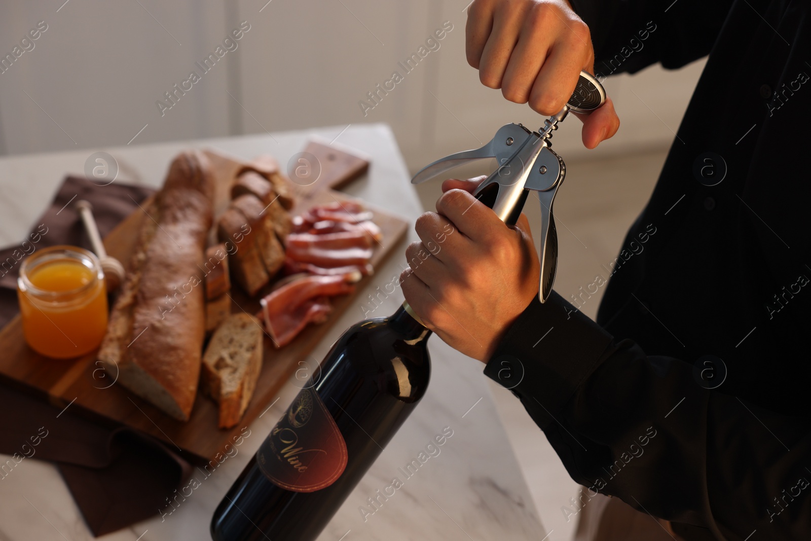 Photo of Romantic dinner. Man opening wine bottle with corkscrew at table in kitchen, closeup