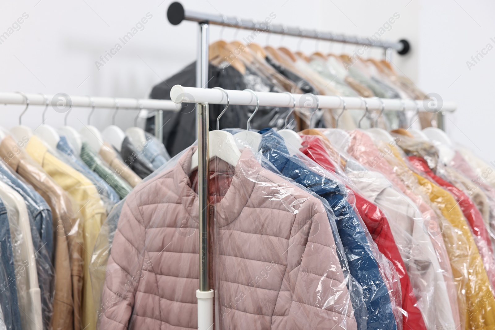 Photo of Dry-cleaning service. Many different clothes in plastic bags hanging on rack indoors, closeup