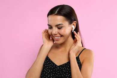 Happy young woman listening to music through wireless earphones on pink background