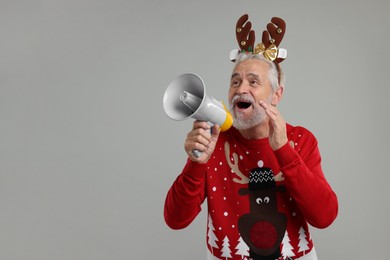 Photo of Senior man in Christmas sweater and reindeer headband shouting in megaphone on grey background. Space for text