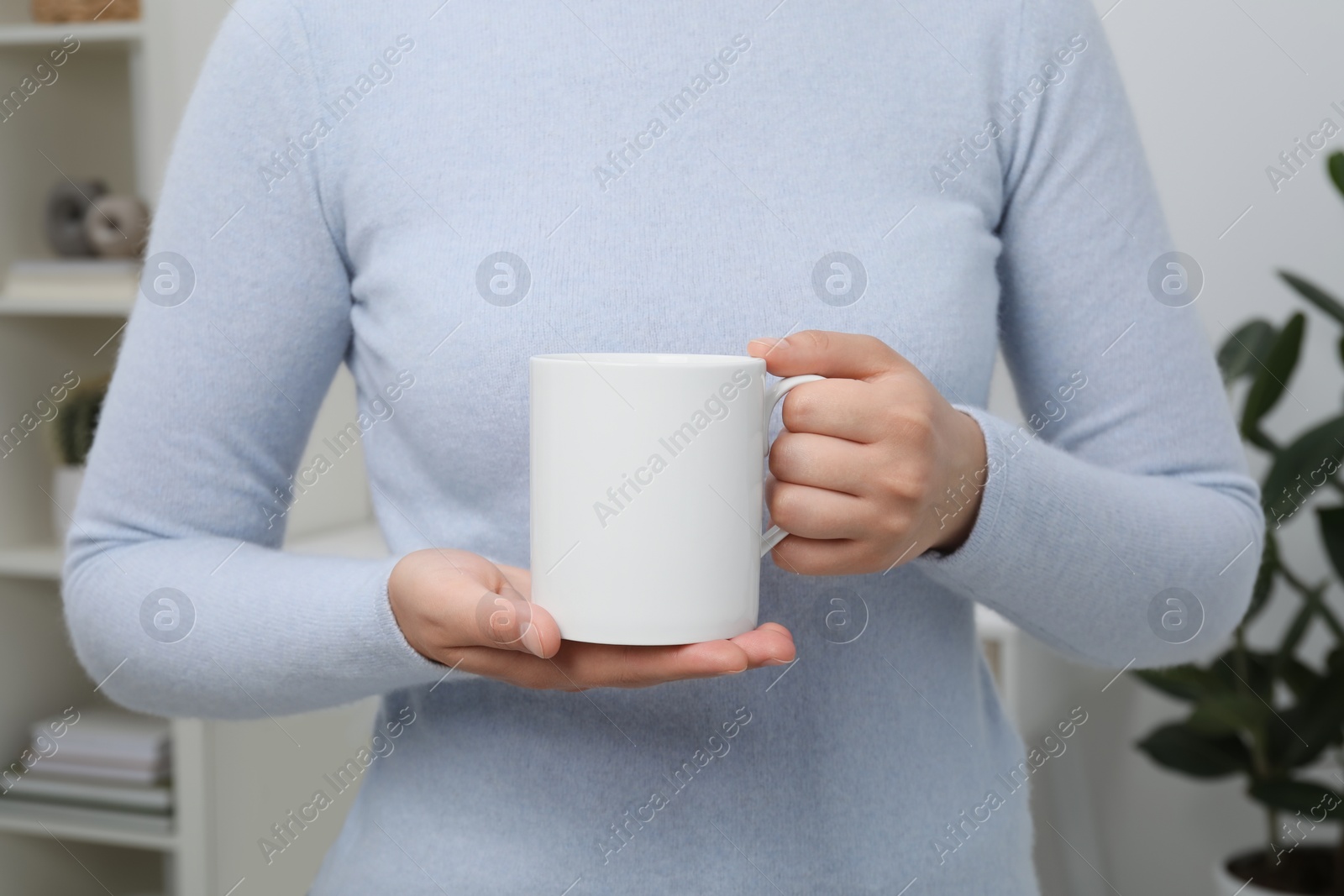 Photo of Woman holding white mug at home, closeup. Mockup for design