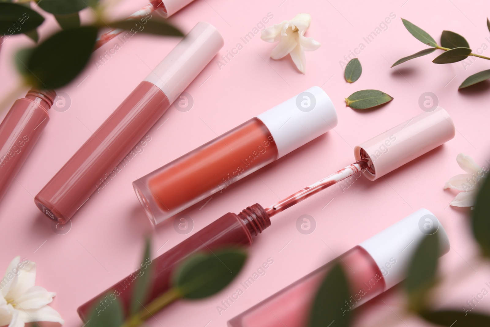Photo of Different lip glosses, applicator, flowers and green leaves on pink background