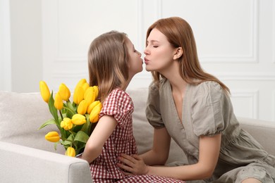 Photo of Cute daughter hiding bouquet of yellow tulips for her mother on sofa at home