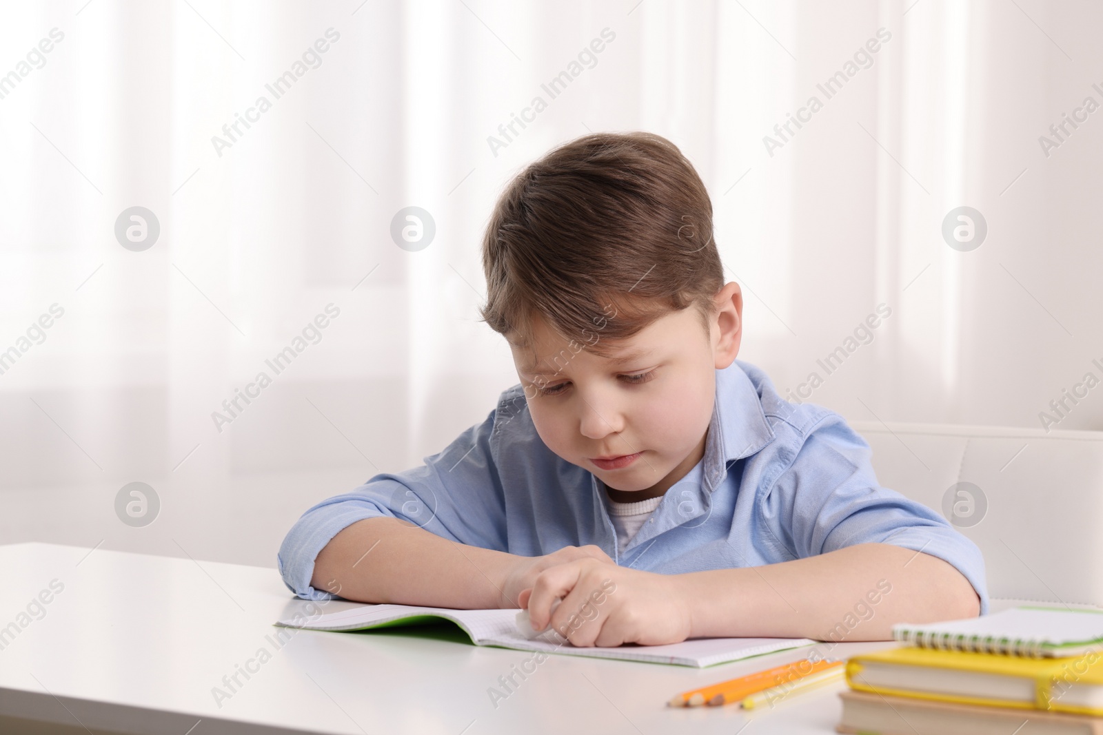 Photo of Little boy erasing mistake in his notebook at white desk indoors