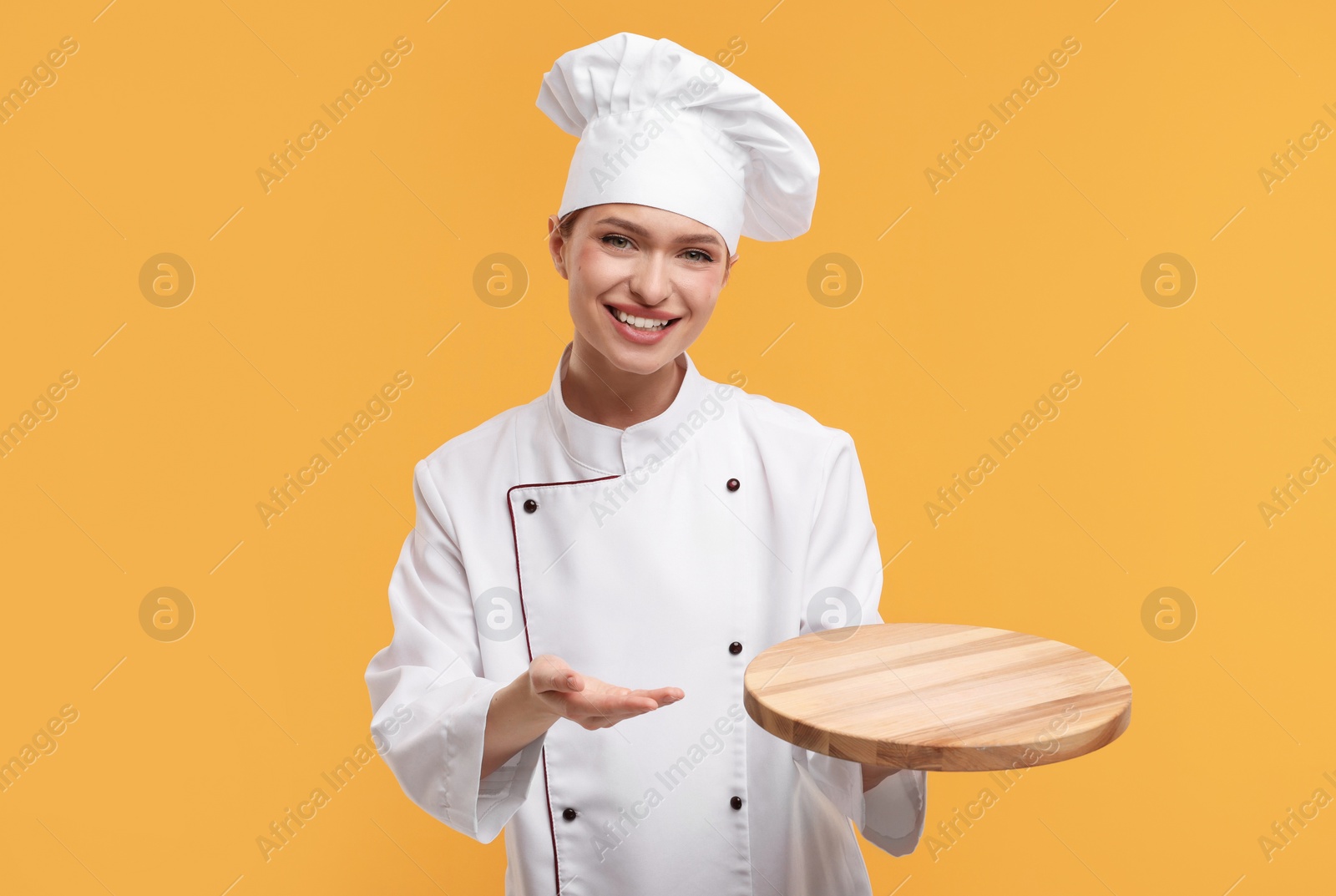 Photo of Happy chef in uniform holding empty wooden board on orange background