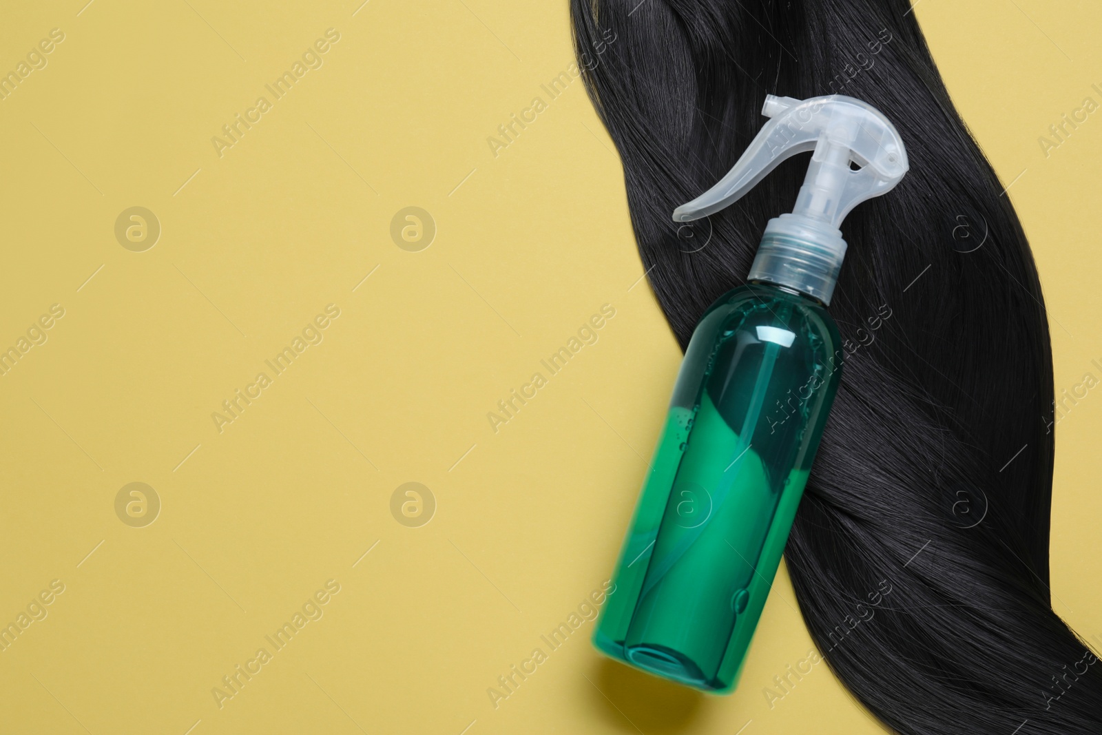 Photo of Spray bottle with thermal protection and lock of black hair on yellow background, flat lay. Space for text
