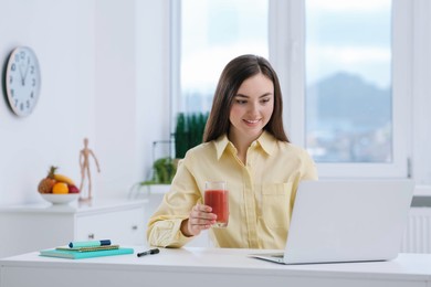 Photo of Beautiful young woman with delicious smoothie using laptop at table in office