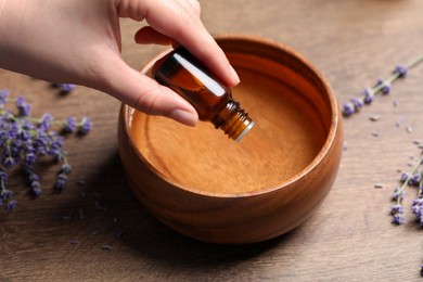 Woman dripping essential oil from bottle into bowl near lavender at wooden table, closeup