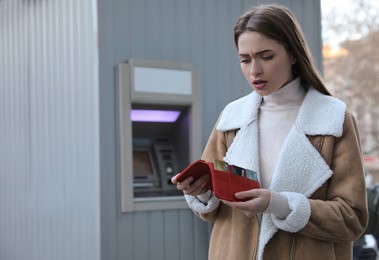 Unhappy young woman with wallet near cash machine outdoors