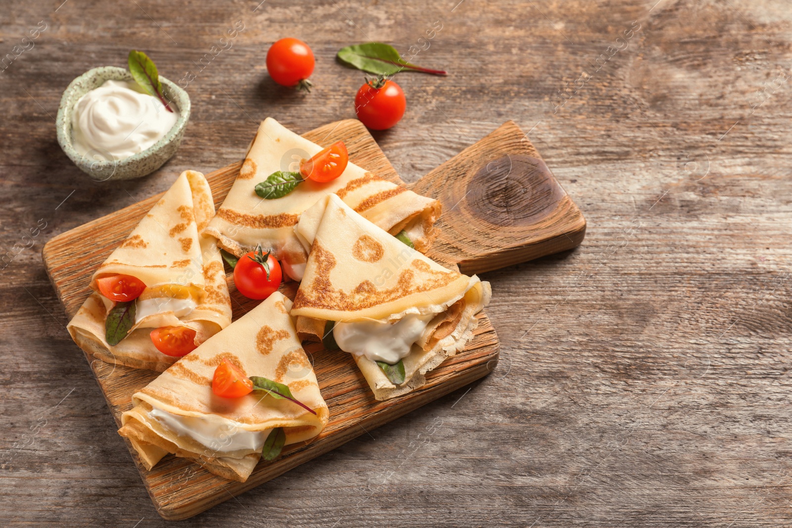 Photo of Thin pancakes with sour cream and cherry tomatoes on wooden board