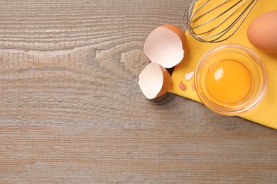 Raw chicken egg, bowl with yolk and whisk on wooden table, flat lay. Space for text