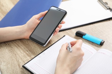 Photo of Woman taking notes while using smartphone at wooden table, closeup