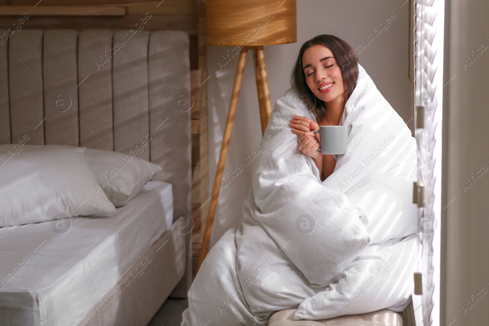 Photo of Beautiful young woman wrapped with soft blanket enjoying hot drink near window at home. Space for text