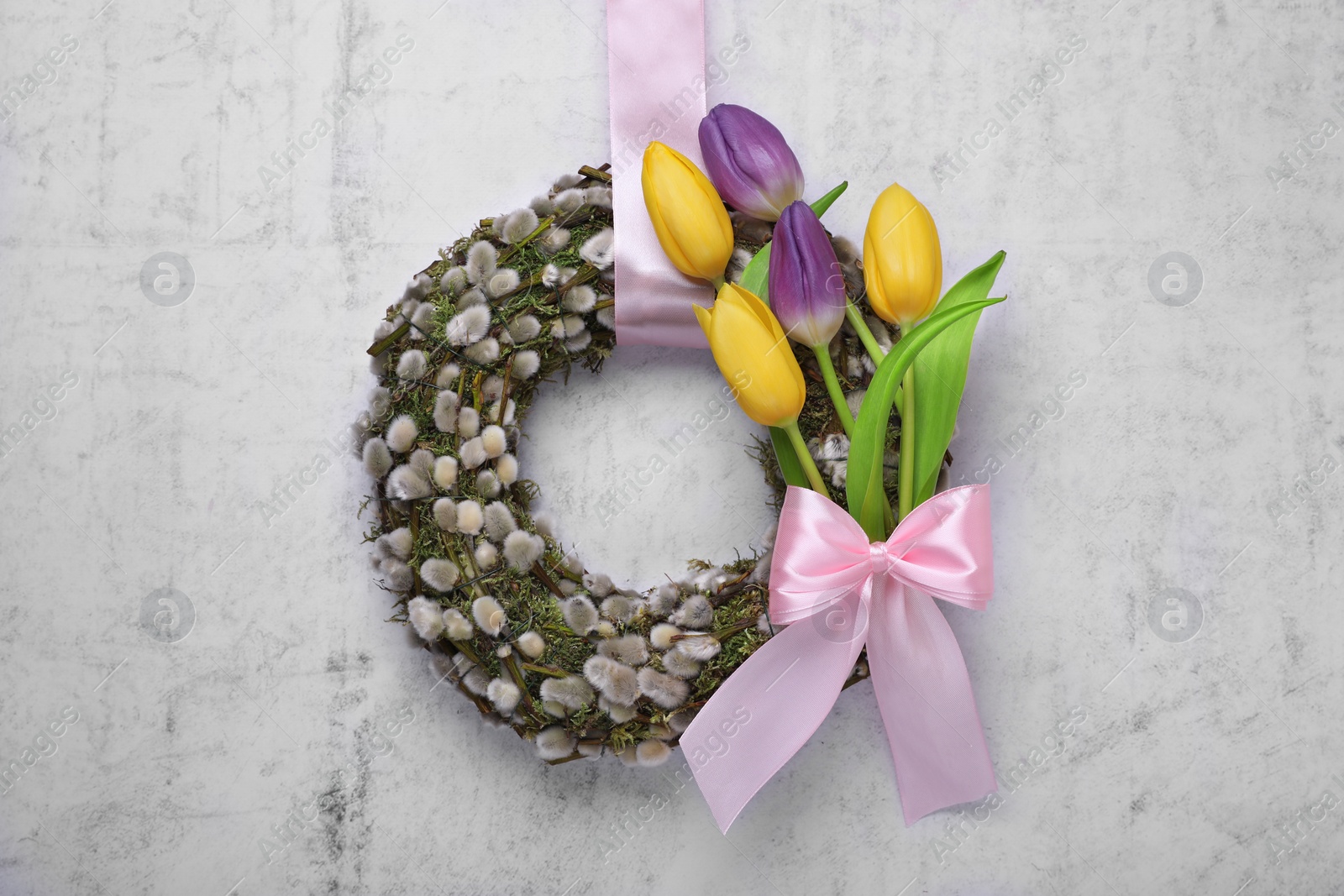 Photo of Wreath made of beautiful willow, colorful tulip flowers and pink bow hanging on light background