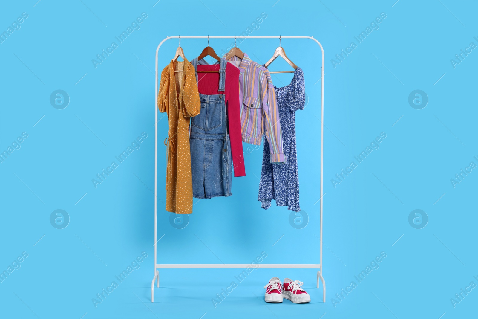 Photo of Rack with stylish women`s clothes on wooden hangers and sneakers against light blue background