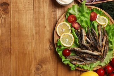 Photo of Delicious fried anchovies served with lemon, tomatoes and sauce on wooden table, flat lay. Space for text