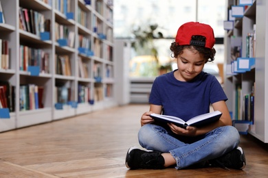 Photo of Cute little boy reading book on floor in library