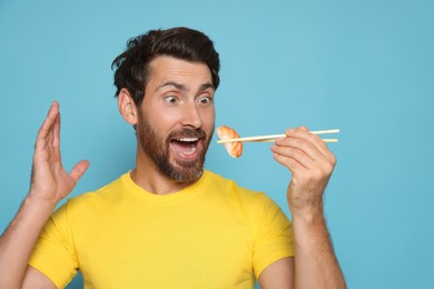 Emotional man holding tasty sushi with chopsticks on light blue background