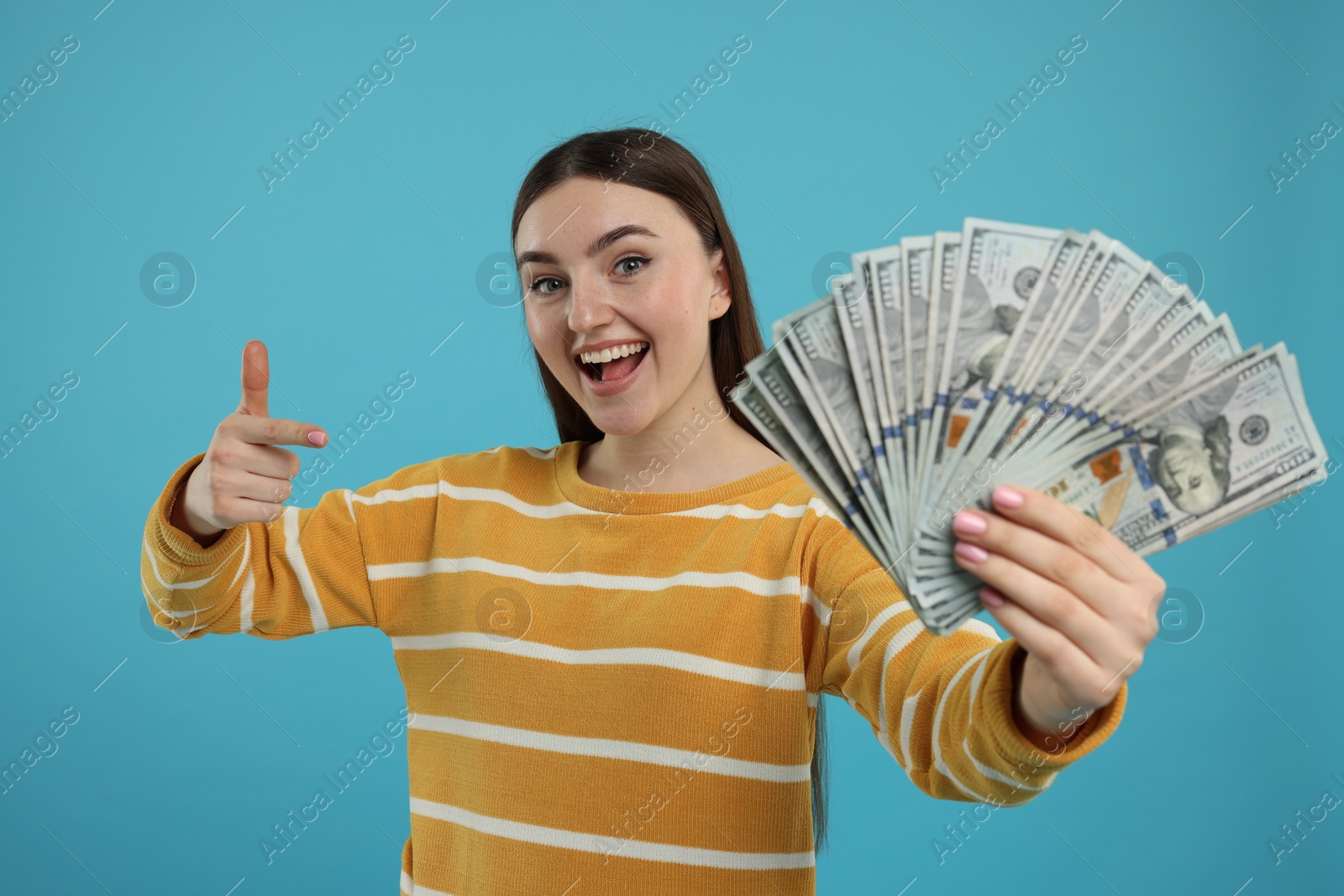 Photo of Happy woman pointing at dollar banknotes on light blue background, selective focus