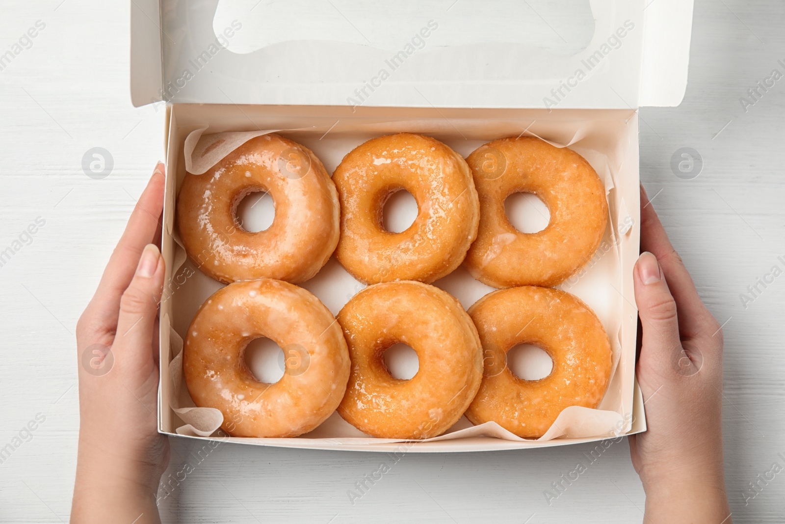 Photo of Woman with box of delicious donuts at white wooden table, top view