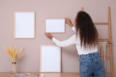 Photo of African American woman hanging empty frame on pale rose wall over table in room, back view. Mockup for design