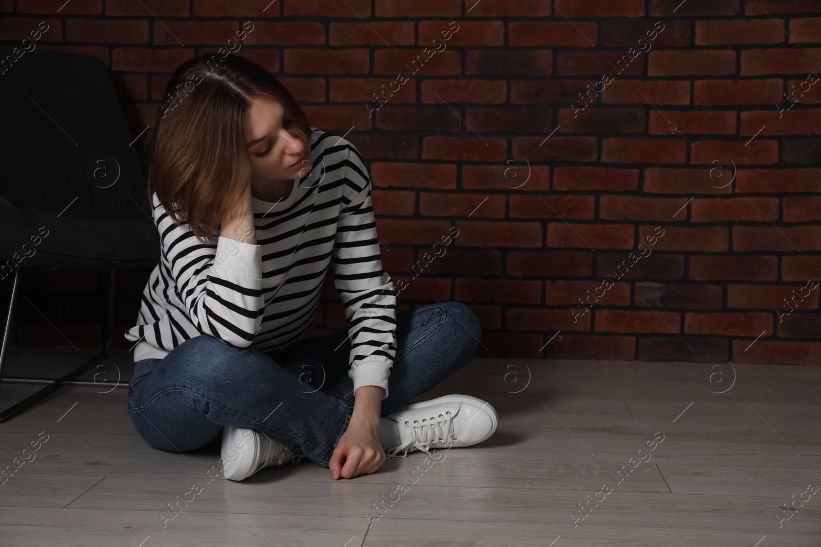 Photo of Sad young woman sitting on floor near brick wall indoors