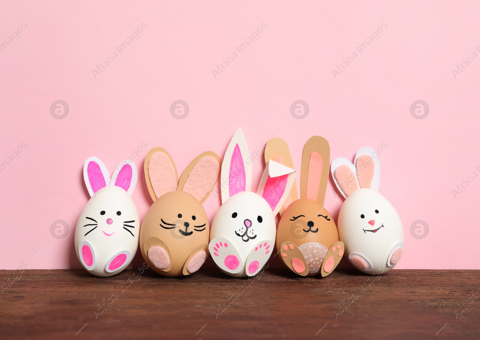 Photo of Eggs as cute bunnies on wooden table against pink background. Easter celebration