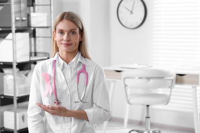 Doctor with stethoscope and pink ribbon indoors, space for text. Breast cancer awareness