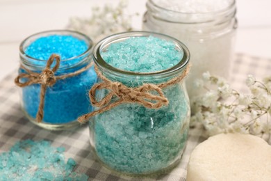 Jars with different sea salt and flowers on towel, closeup