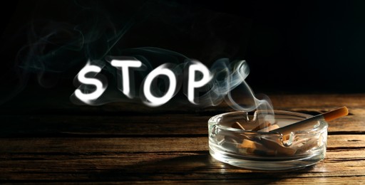 Image of Quit smoking. Word Stop of smoke, ashtray with stubs and smoldering cigarette on wooden table against black background, banner design