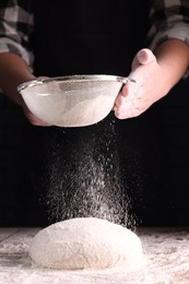 Photo of Man sprinkling flour over dough at wooden table on dark background, closeup