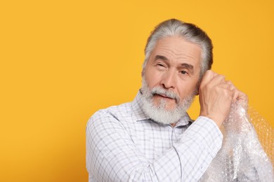 Emotional senior man popping bubble wrap on yellow background, space for text. Stress relief