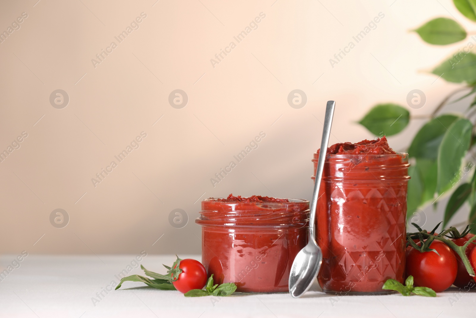 Photo of Jars of tasty tomato paste, spoon and ingredients on white table, space for text