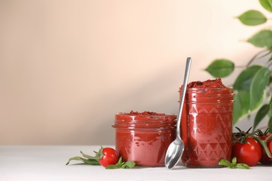 Photo of Jars of tasty tomato paste, spoon and ingredients on white table, space for text
