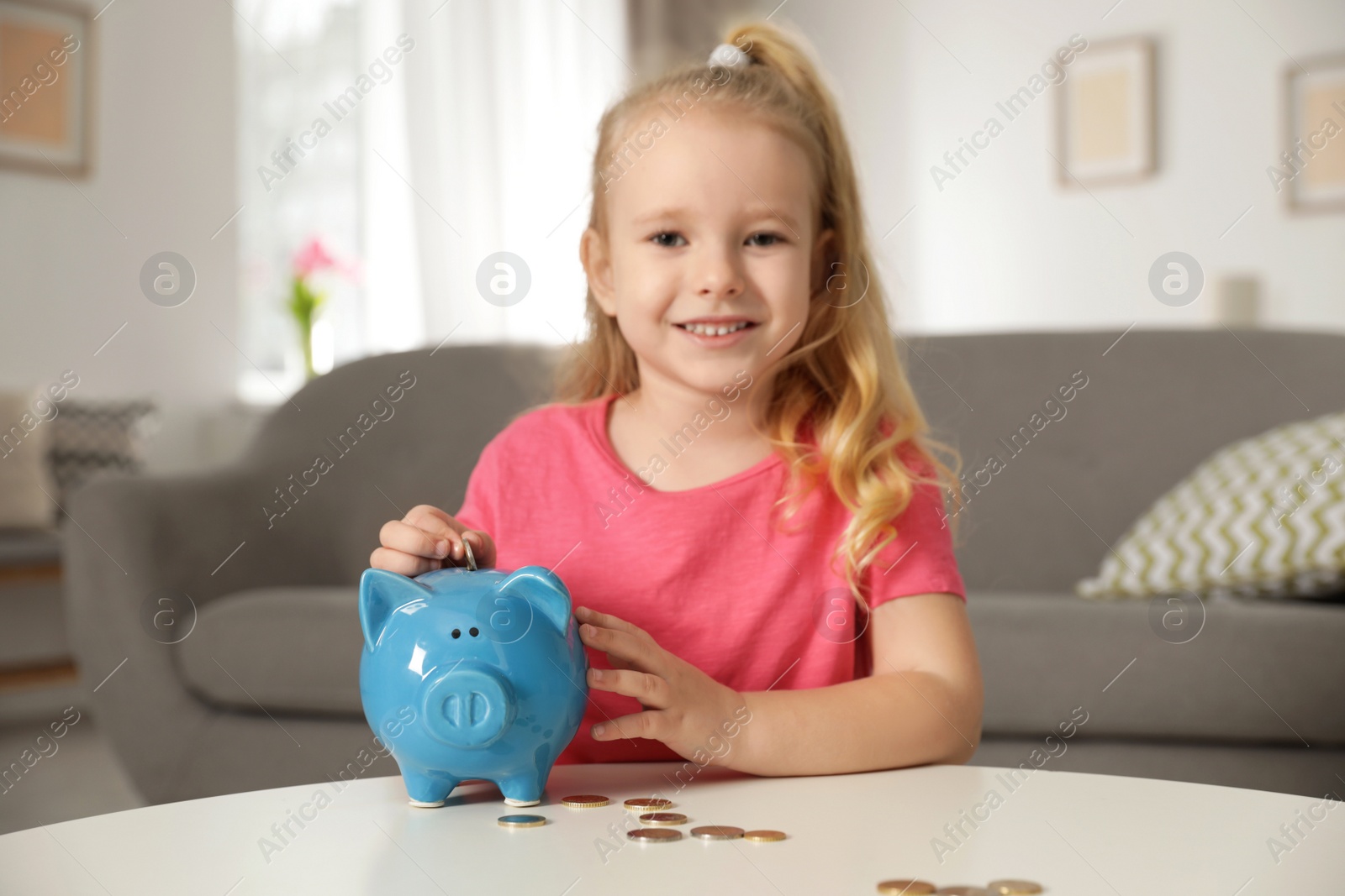 Photo of Cute girl putting coin into piggy bank at table in living room. Saving money