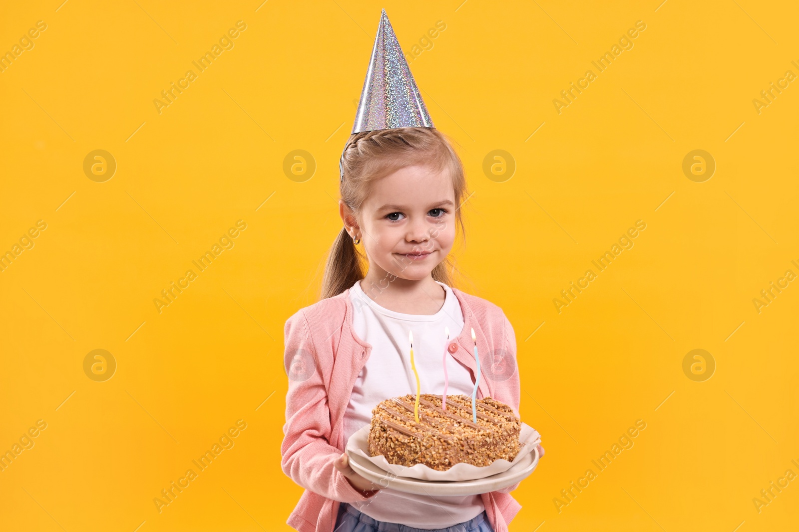 Photo of Birthday celebration. Cute little girl in party hat holding tasty cake with burning candles on orange background