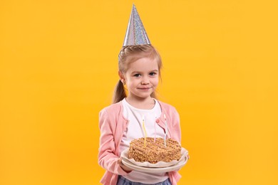 Birthday celebration. Cute little girl in party hat holding tasty cake with burning candles on orange background