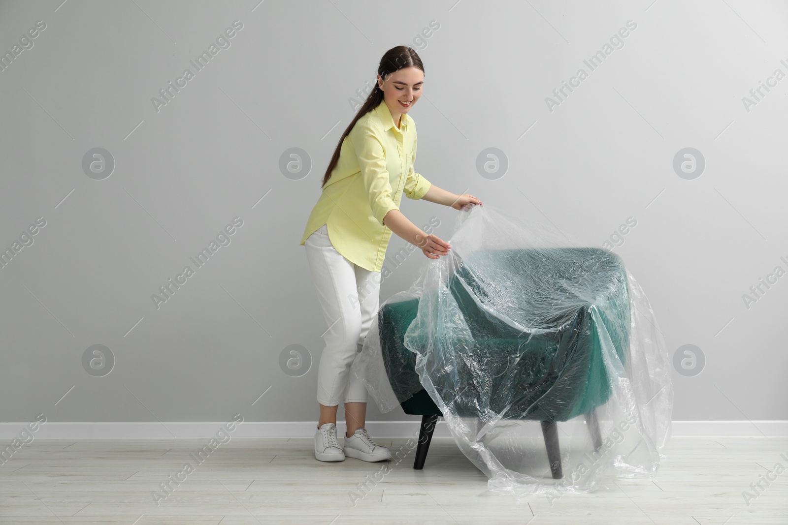 Photo of Young woman putting plastic film away from armchair near light grey wall indoors
